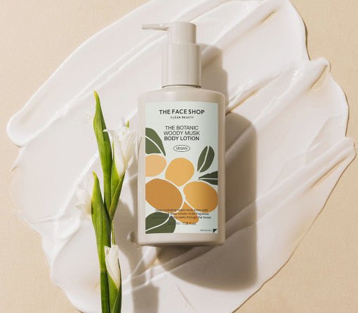 Pump bottle of woody musk body lotion on creamy white texture with green leaves and white flower beside it