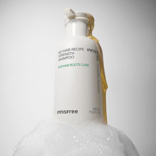 White shampoo bottle with golden liquid pouring down side, surrounded by foam bubbles on gray background