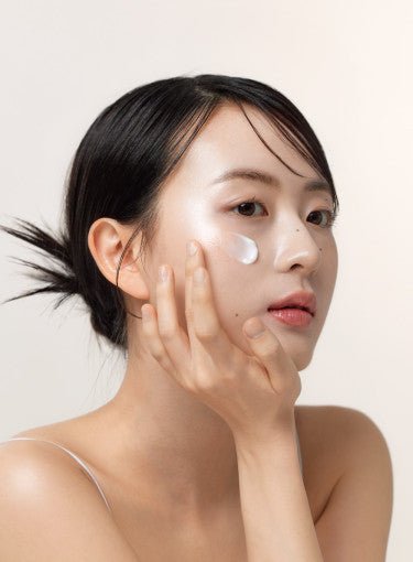 Young woman with smooth skin applying hydrating cream to cheek against neutral background