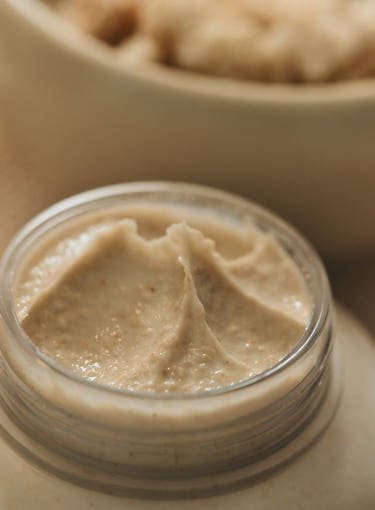 Creamy beige mask texture with fine grainy particles in an open jar suggesting gentle radiant skin treatment