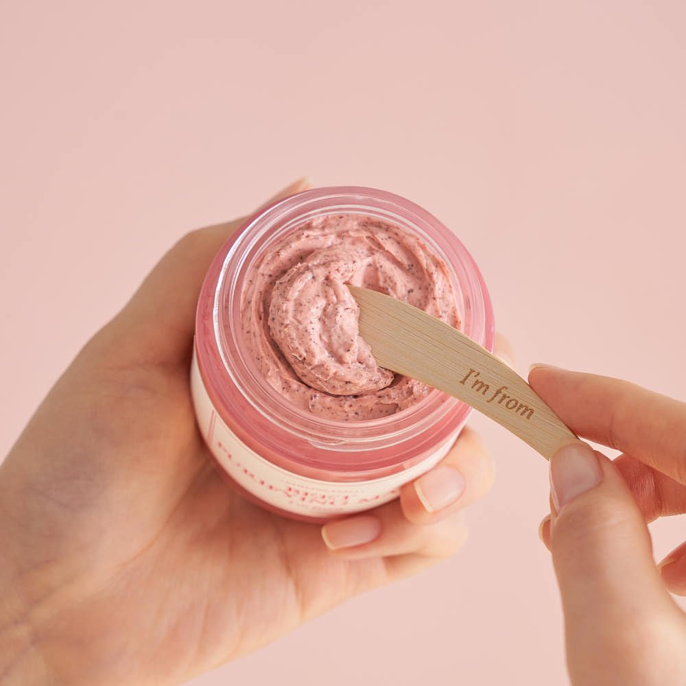 Hands holding pink purifying mask cream with wooden spatula against soft pink background