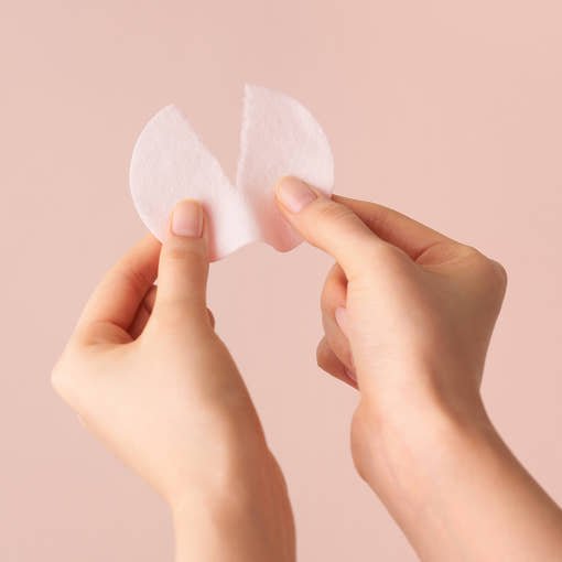 Hands holding and gently pulling apart a thin white cotton refresh pad against a soft beige background