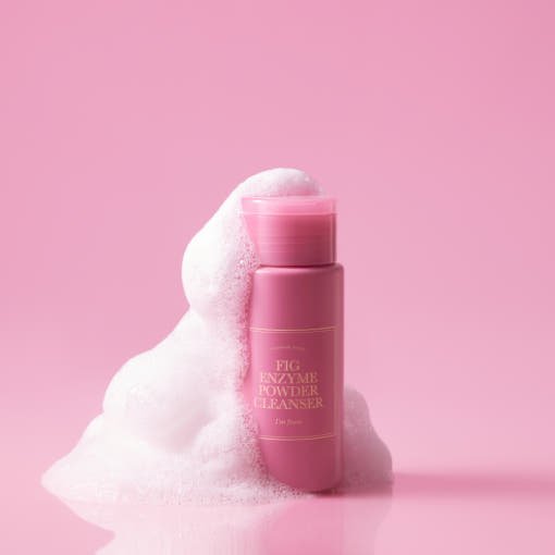 Pink bottle of enzyme powder cleanser surrounded by white foamy bubbles on a solid pink background