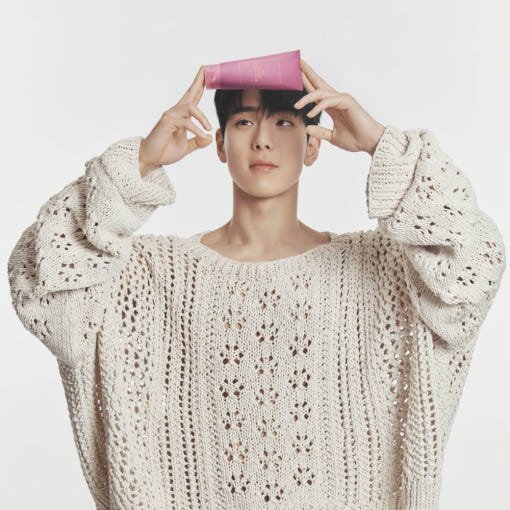 Young man in a cream knitted sweater holding a pink cleanser tube above his head with a neutral background