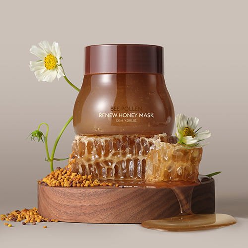 Amber jar of bee pollen mask with honeycomb pieces, pollen granules, and white flowers on wooden platform