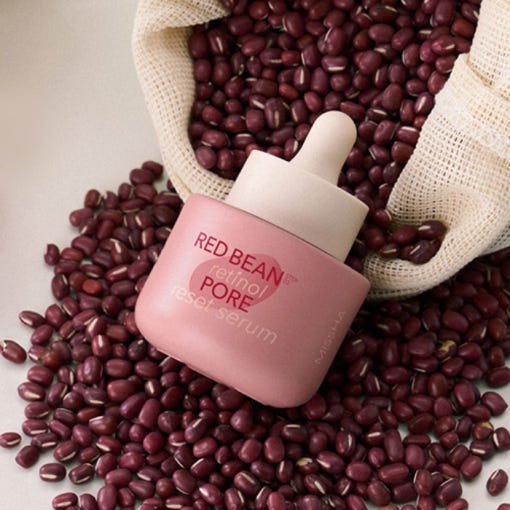 Pink dropper bottle of pore minimizing serum surrounded by red beans spilling from a woven sack