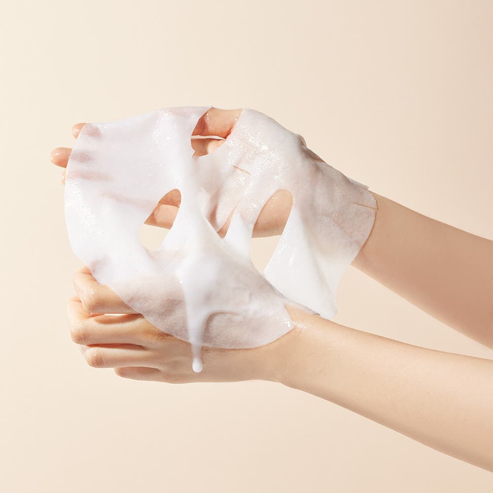 Hands holding a moist, translucent sheet mask with a creamy texture against a soft beige background