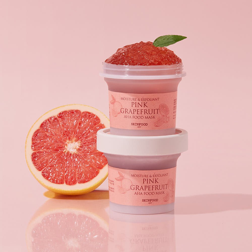 Two stacked jars of pink grapefruit exfoliating mask with fresh grapefruit half and mint leaf on pink backdrop
