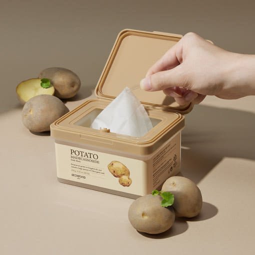 Hand pulling a sheet mask from a beige container labeled with potato illustration surrounded by fresh potatoes with green sprouts