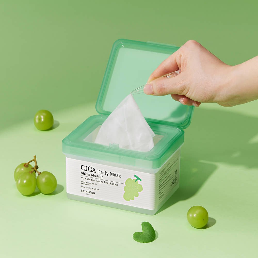Hand lifting a sheet mask with tweezers from a green-topped container surrounded by Shine Muscat grapes on green background