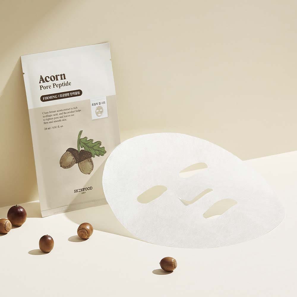 Sheet mask lying flat next to beige acorn-themed pouch with small acorns scattered on light background