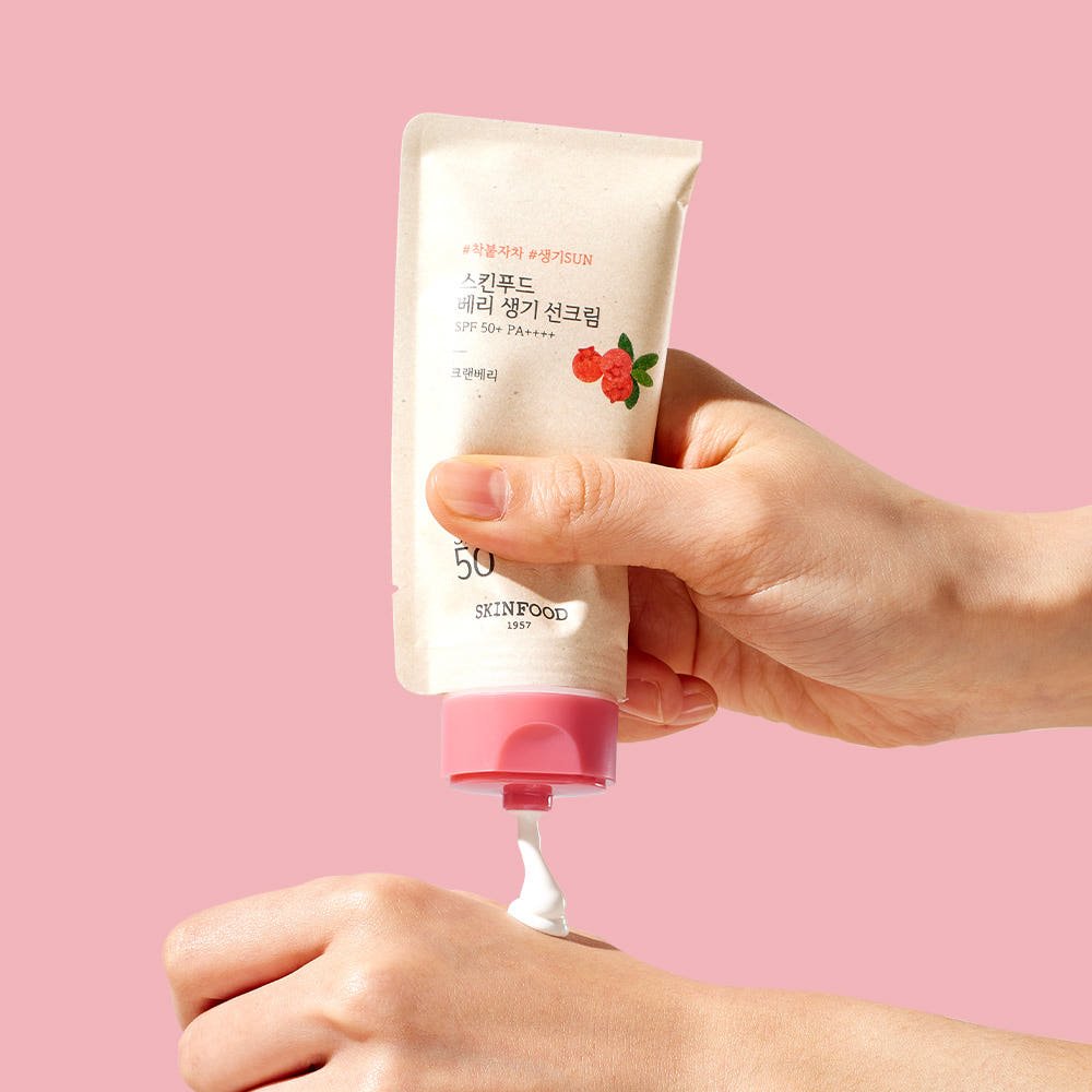 Hand squeezing white berry glowing sun cream with SPF50+ from beige tube with pink cap onto wrist on pink background