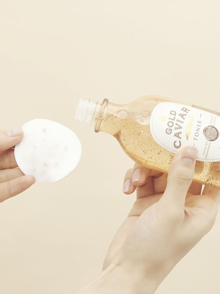 Hand pouring golden toner liquid from clear bottle onto white cotton pad against beige background