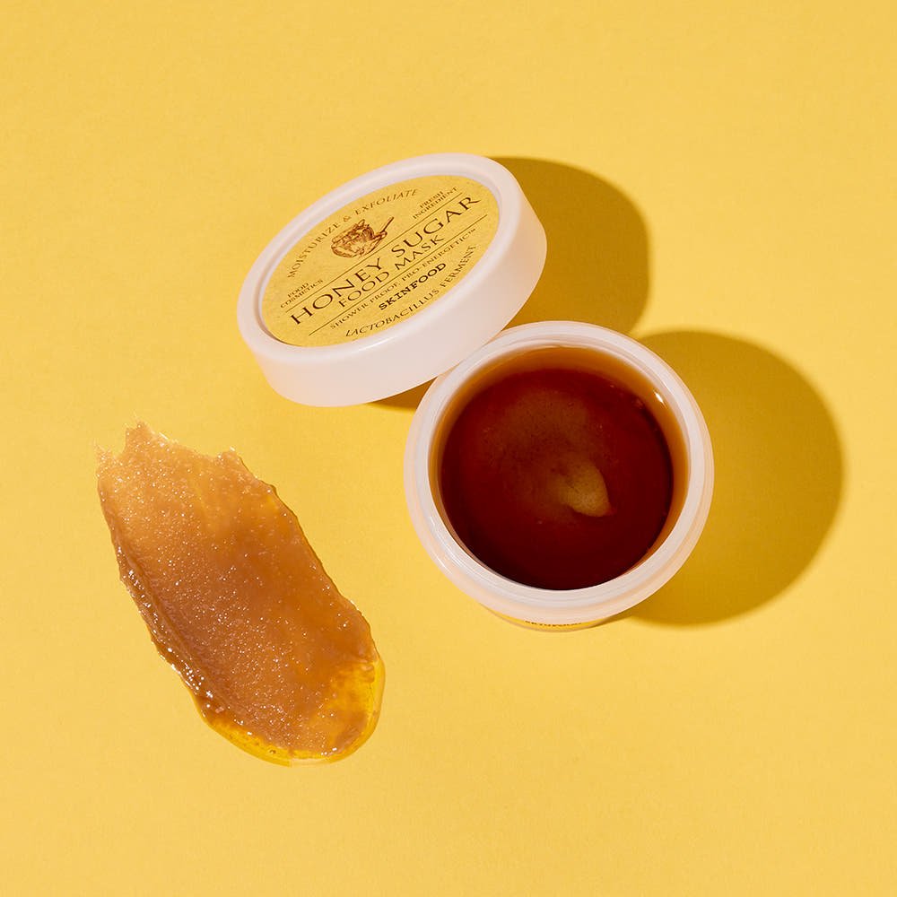 Open jar of honey-colored exfoliating sugar mask with smooth texture beside a smear on yellow background