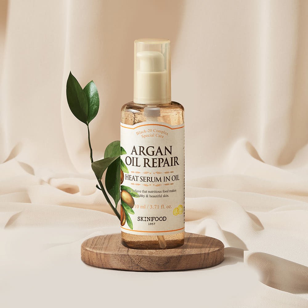 SKINFOOD Argan Oil Repair Plus Heat Serum in Oil – 110ml pump bottle on wooden platform with green leaf.