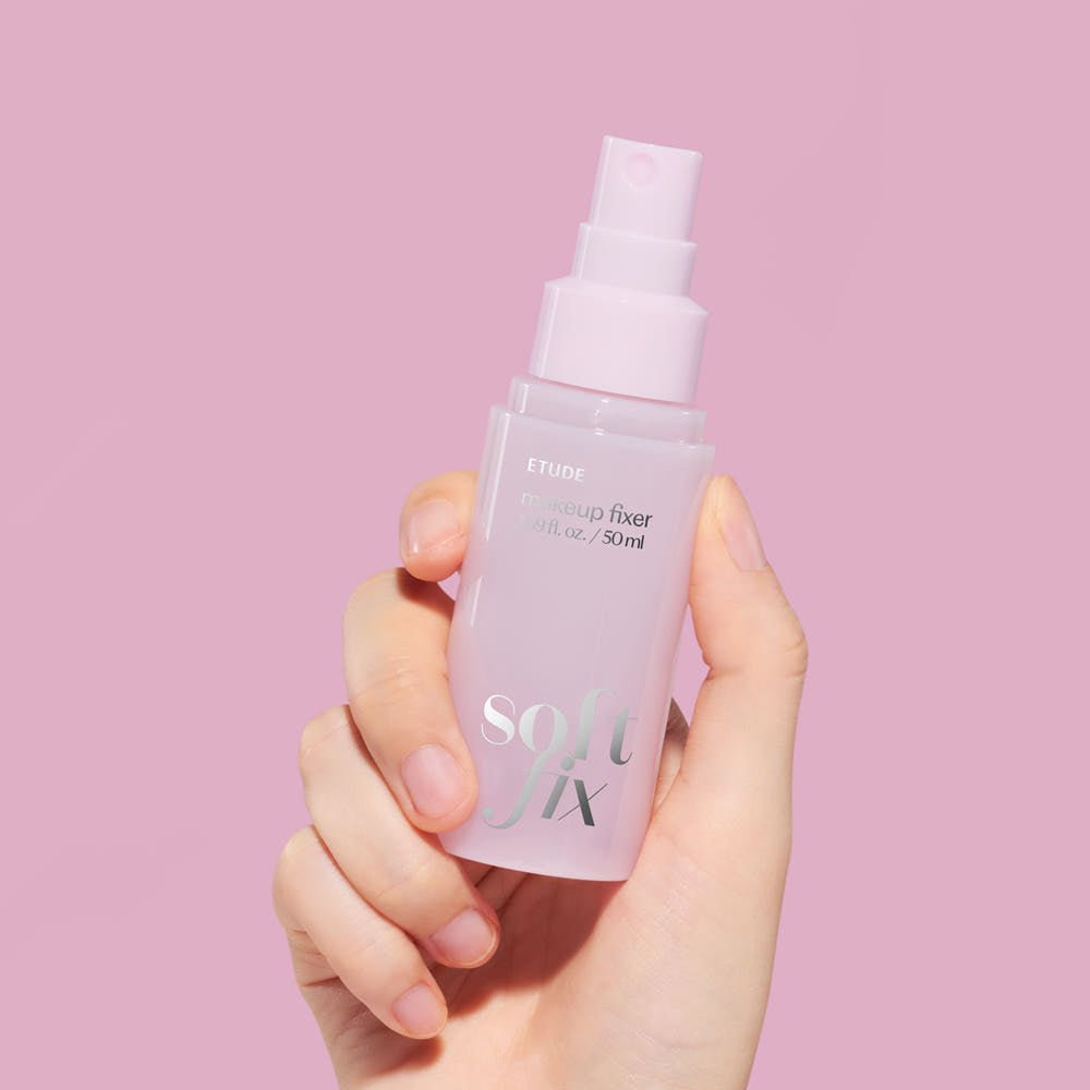 Hand holding ETUDE Soft Fix Makeup Fixer 50ml spray bottle with pastel pink background for long-lasting hold