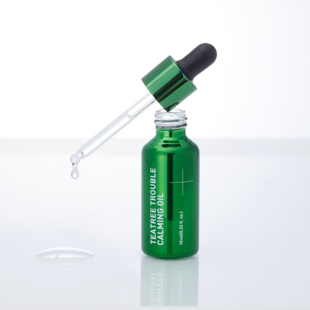 Green glass dropper bottle with clear calming oil and dropper releasing a droplet over reflective surface