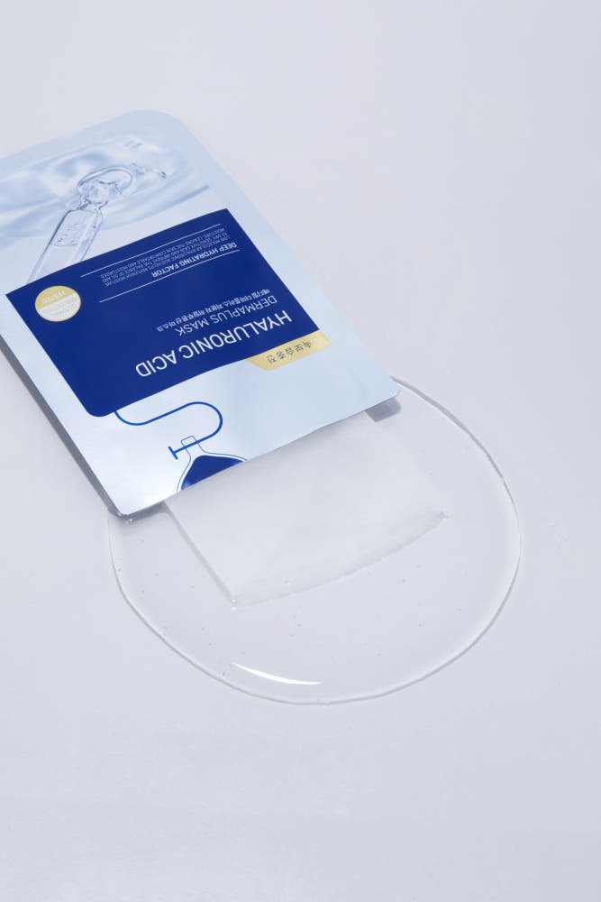 Blue and white hyaluronic acid face mask packet lying on clear gel spilled on white surface