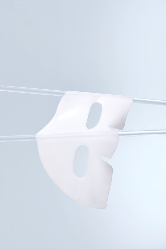 White gel facial sheet mask hanging on thin rods against minimal pale blue background