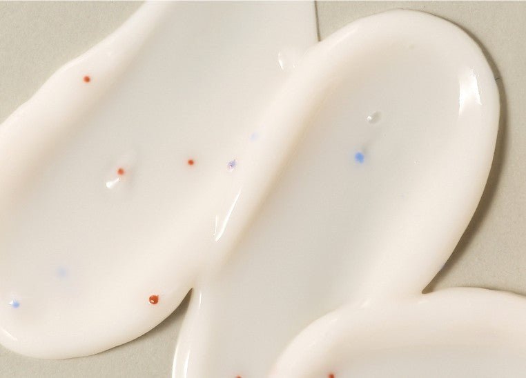 Close-up of creamy white serum with small blue and red exfoliating beads on a light surface