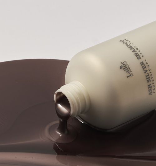 Matte bottle of ash silver shampoo pouring thick, smooth, dark silver-gray liquid onto white surface.