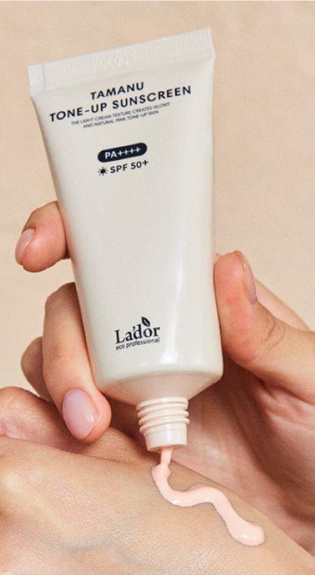 Hand squeezing light pink tinted sunscreen cream from white tube onto back of other hand against neutral background