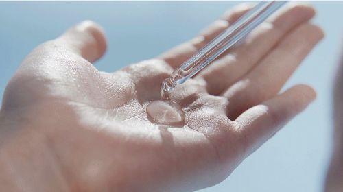 Clear hair oil droplet being dispensed onto an open palm against a blurred blue background