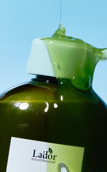 Green gel shampoo pouring over white cap of dark amber bottle against pale blue background