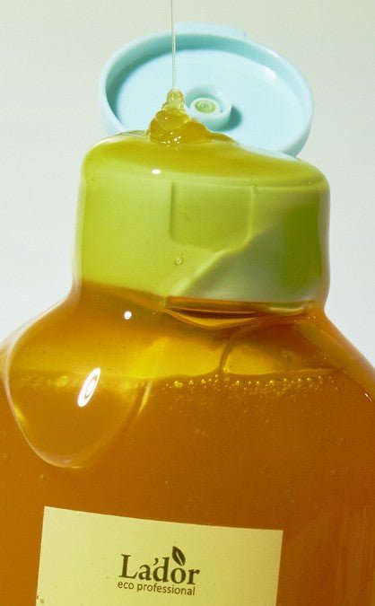 Close-up of golden shampoo liquid being poured from an open pale green cap on amber bottle labeled Lador eco professional