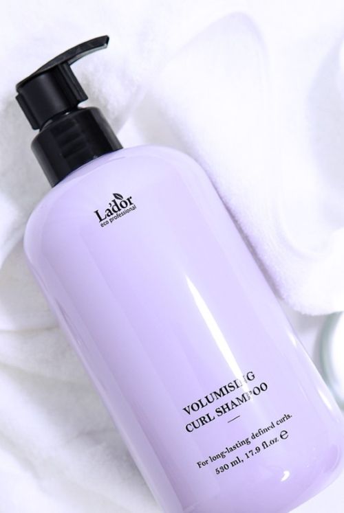 Tall light purple pump bottle of volumising curl shampoo resting on white towels.