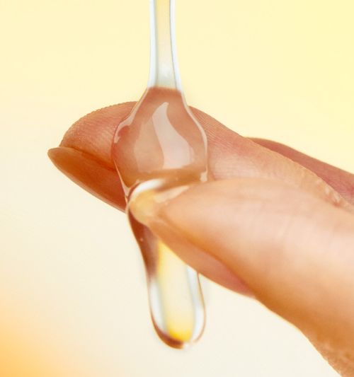 Close-up of a finger holding a clear, nourishing liquid drop against a soft yellow background