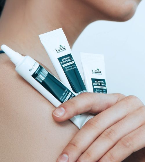Hand holding three tubes of keratin power glue hair treatment against bare shoulder background
