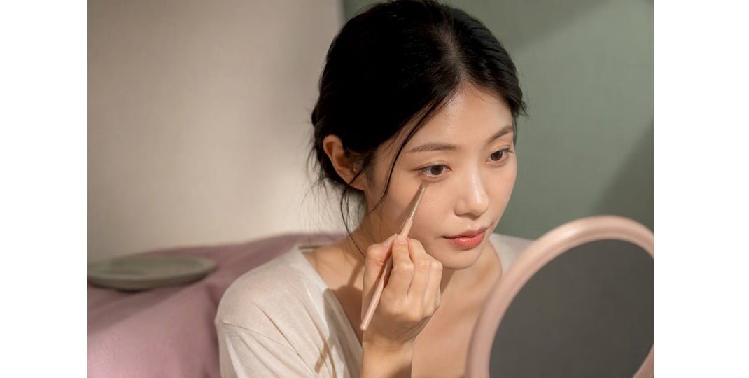 Woman applying eye makeup with a slim brush while looking into a handheld mirror