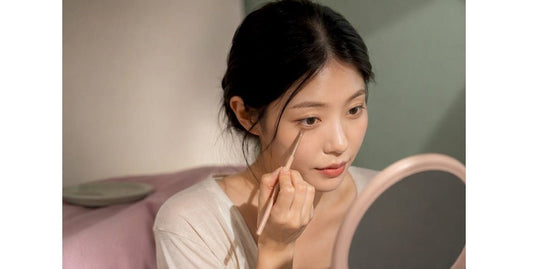 Woman applying eye makeup with a slim brush while looking into a handheld mirror