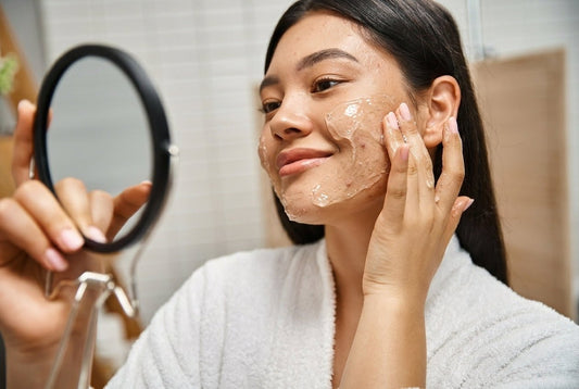 Young woman in a white robe applying a skincare cleanser while looking in a mirror.
