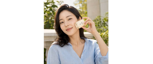 Smiling woman in a light blue blouse holding a small white sunscreen stick beside her face in an outdoor setting