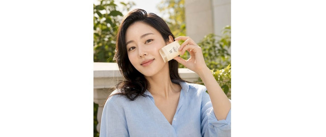 Smiling woman in a light blue blouse holding a small white sunscreen stick beside her face in an outdoor setting