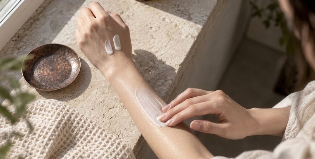 Person spreading white cream across a forearm beside a small bowl on a sunlit stone surface