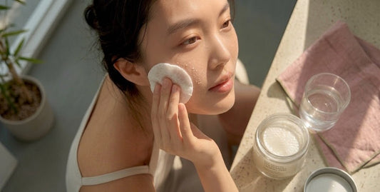 Woman applying a cotton pad to her cheek beside glass jars and a tumbler on a sunlit table