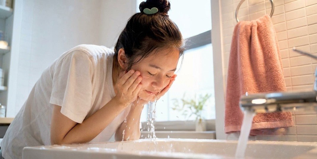 Woman rinsing her face at a bathroom sink with water splashing and a pink towel nearby