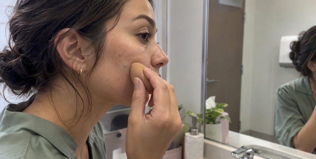 Woman in side profile examining her cheek in a bathroom mirror while gently pressing the skin with her fingertips