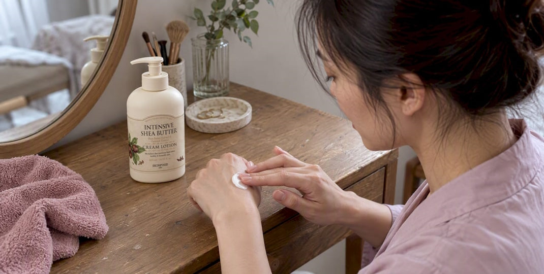 Woman applying cream to her hand beside a white skincare bottle on a wooden vanity in a softly lit bedroom