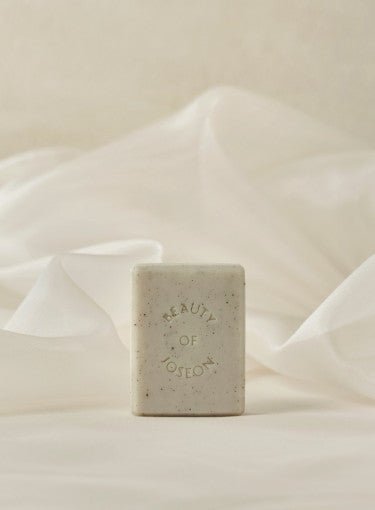 Beige rectangular cleansing bar with subtle speckles and embossed text on soft cream fabric background