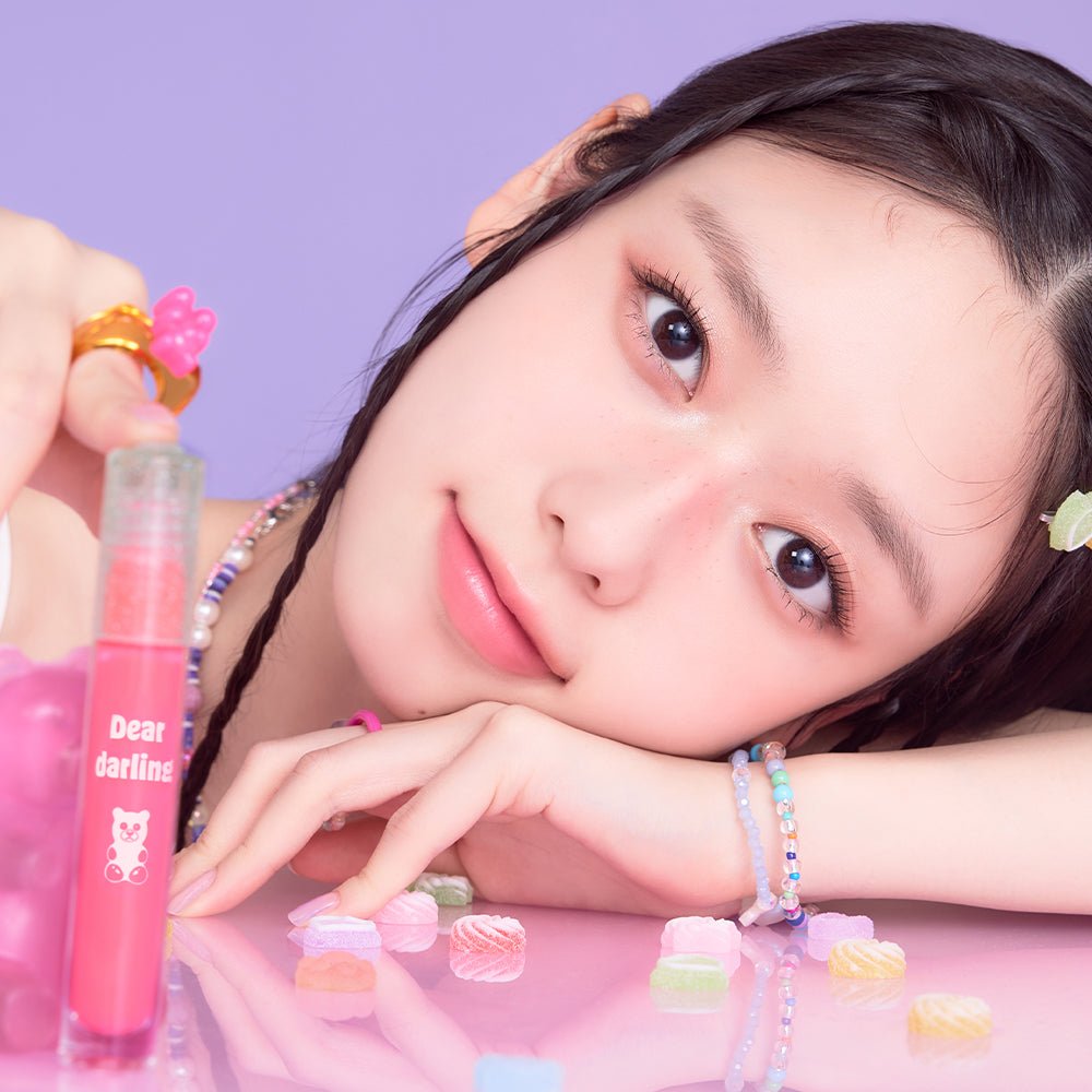 A young woman resting her head on a pink surface holding ETUDE Jelly Pang Dear Darling Water Gel Tint 5g with candy pieces nearby.