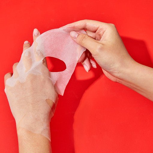 Hands peeling a translucent sheet mask from skin against a vibrant red background showing soothing skincare use