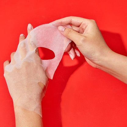 Hands peeling a translucent sheet mask from skin against a vibrant red background showing soothing skincare use