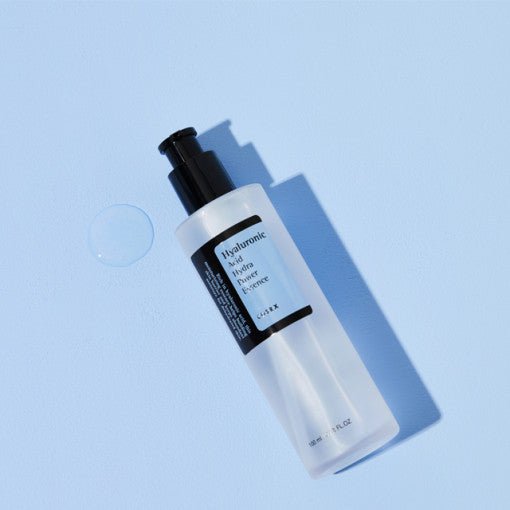 Clear pump bottle of hydrating essence on pale blue surface with a drop of clear serum nearby