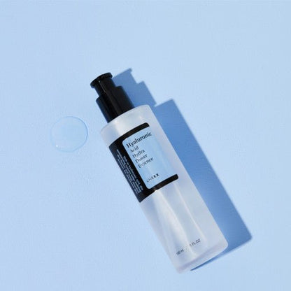 Clear pump bottle of hydrating essence on pale blue surface with a drop of clear serum nearby