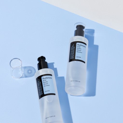 Two translucent bottles of hydrating essence with black pump tops on a pale blue surface casting soft shadows