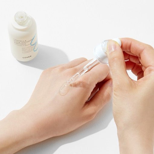Clear hydrating ampoule drop being applied on back of hand with open white bottle nearby on white surface